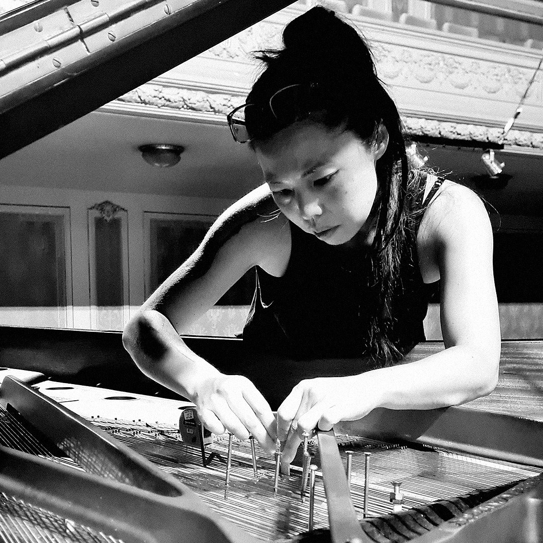 Fotografia em preto e branco. Lilian está em frente a um piano com a tampa aberta. Vê-se do peito para cima. Ela coloca pregos entre as cordas do piano com as duas mãos. Ela é uma mulher de olhos puxados, com pele clara e cabelos longos escuros. Os cabelos estão presos e ela usa óculos de grau sob a cabeça. Usa uma camiseta escura sem mangas.